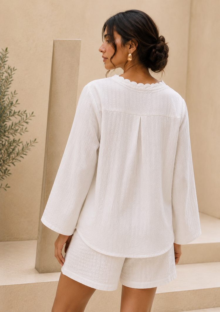 A woman shown from the back wearing a white textured long-sleeve top with a scalloped neckline and matching white shorts, set against a warm beige architectural background.