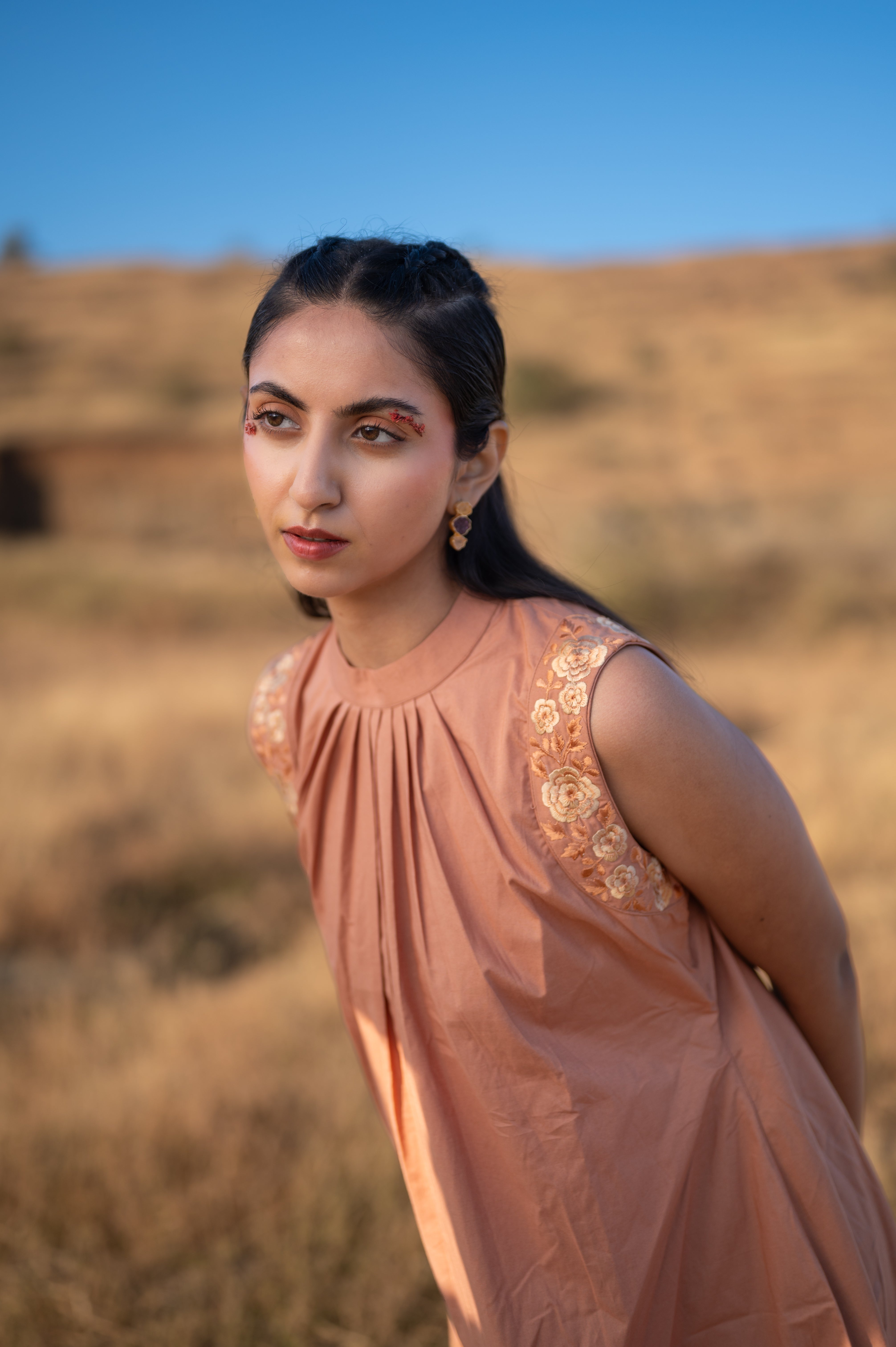 Dessert Rose - Woman in a peach dress with floral embroidery on shoulders standing in a field with a blue sky.