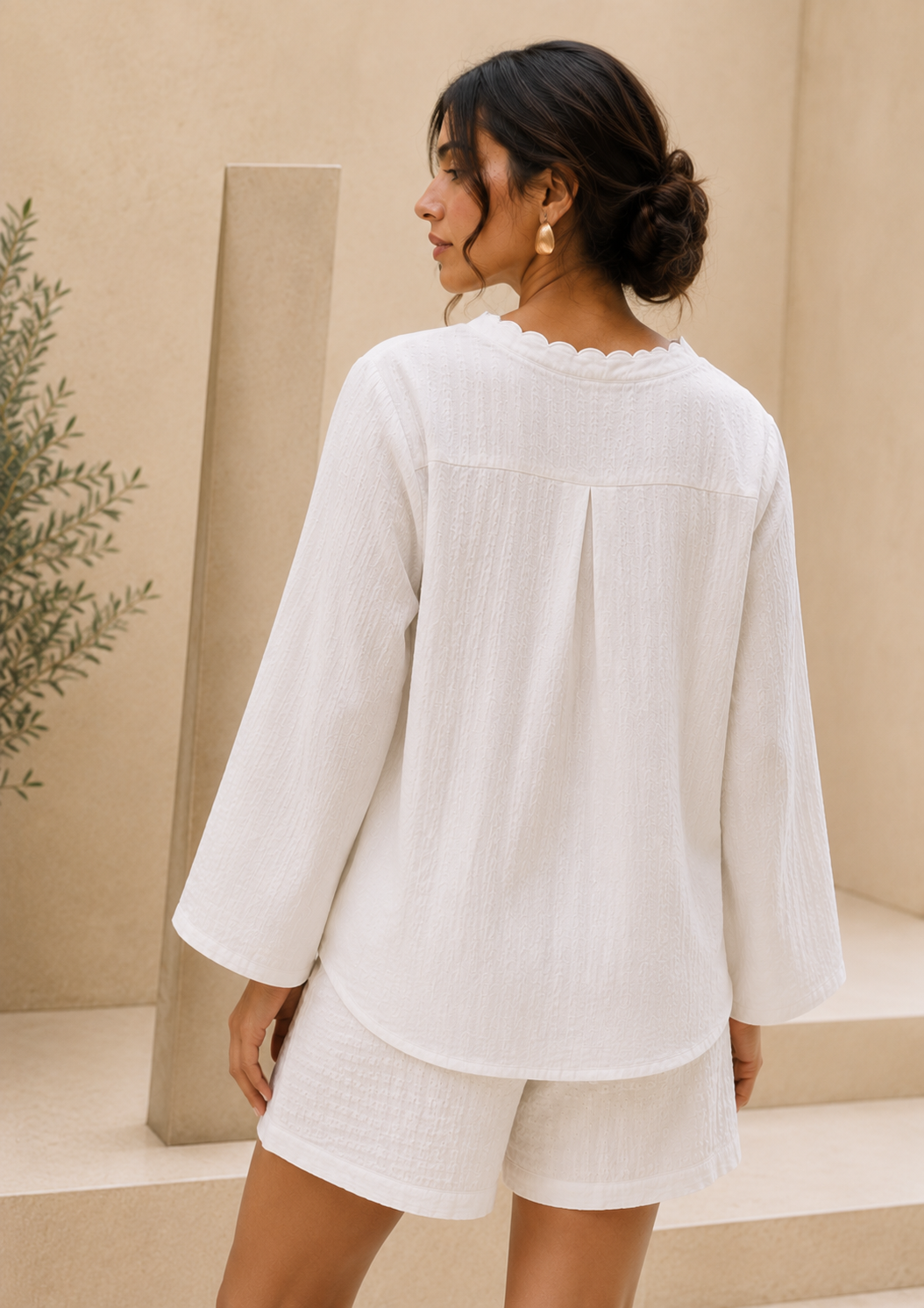 A woman shown from the back wearing a white textured long-sleeve top with a scalloped neckline and matching white shorts, set against a warm beige architectural background.