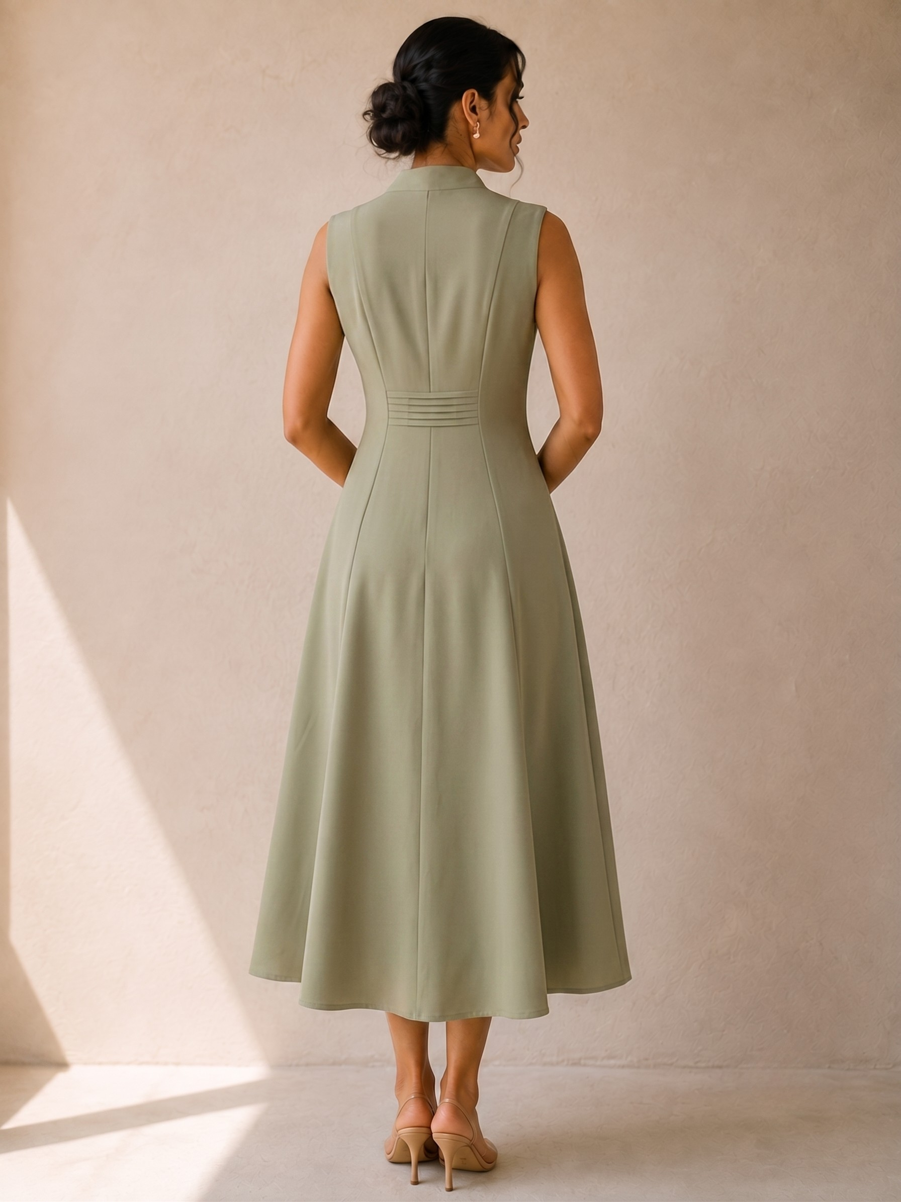 A woman stands with her back to the camera wearing a cords theory sleeveless, high-neck, A-line midi dress in sage green. The dress features structured shoulders, a defined waist with subtle horizontal pleating, and a flared skirt.
