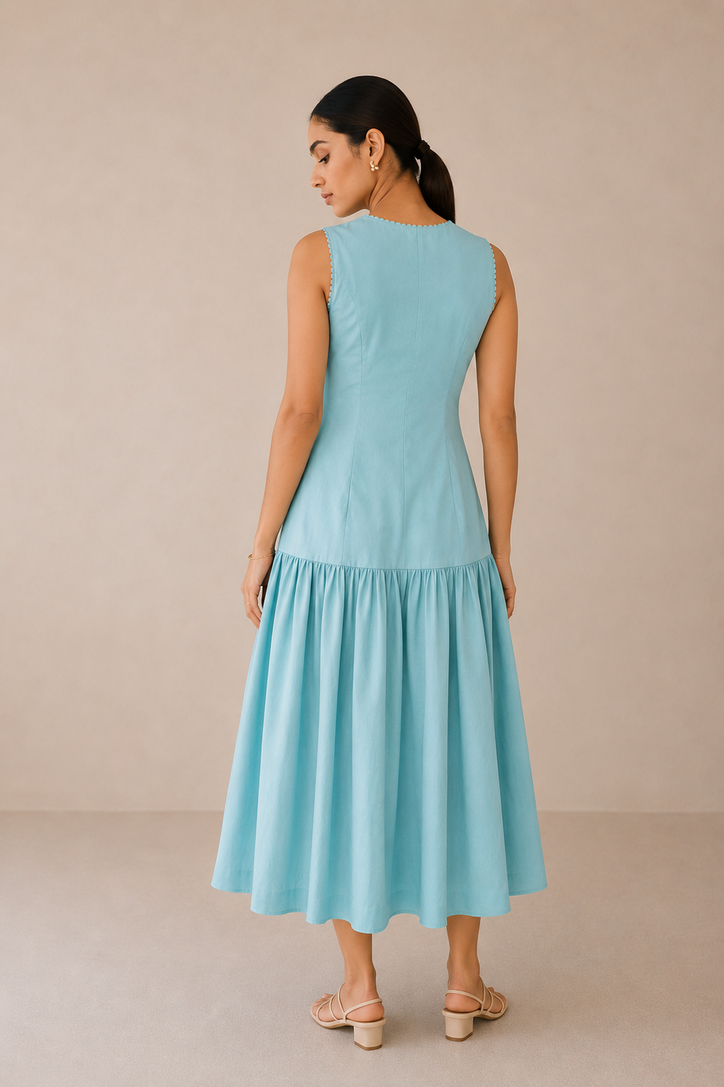 A woman wearing a cords theory sleeveless light blue midi dress with a tiered skirt, shown from the back against a neutral beige studio backdrop.