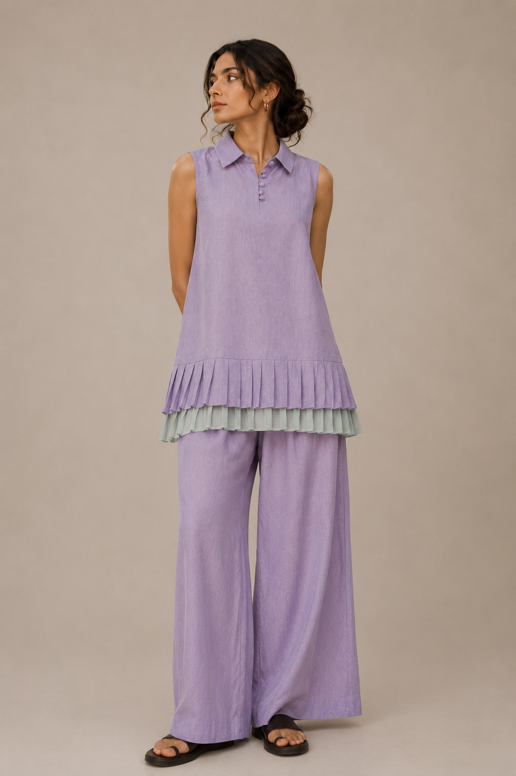 A woman modeling a lavender sleeveless collared top with a ruffled hem and matching wide-leg pants from cords theory, photographed against a plain beige studio background.