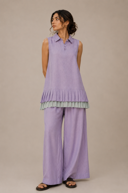 A woman modeling a lavender sleeveless collared top with a ruffled hem and matching wide-leg pants from cords theory, photographed against a plain beige studio background.
