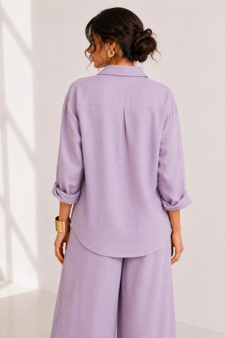 A woman shown from the back wearing a lavender purple matching set consisting of a long-sleeve collared shirt with rolled cuffs and wide-leg trousers, set against a bright white background with soft window shadows.