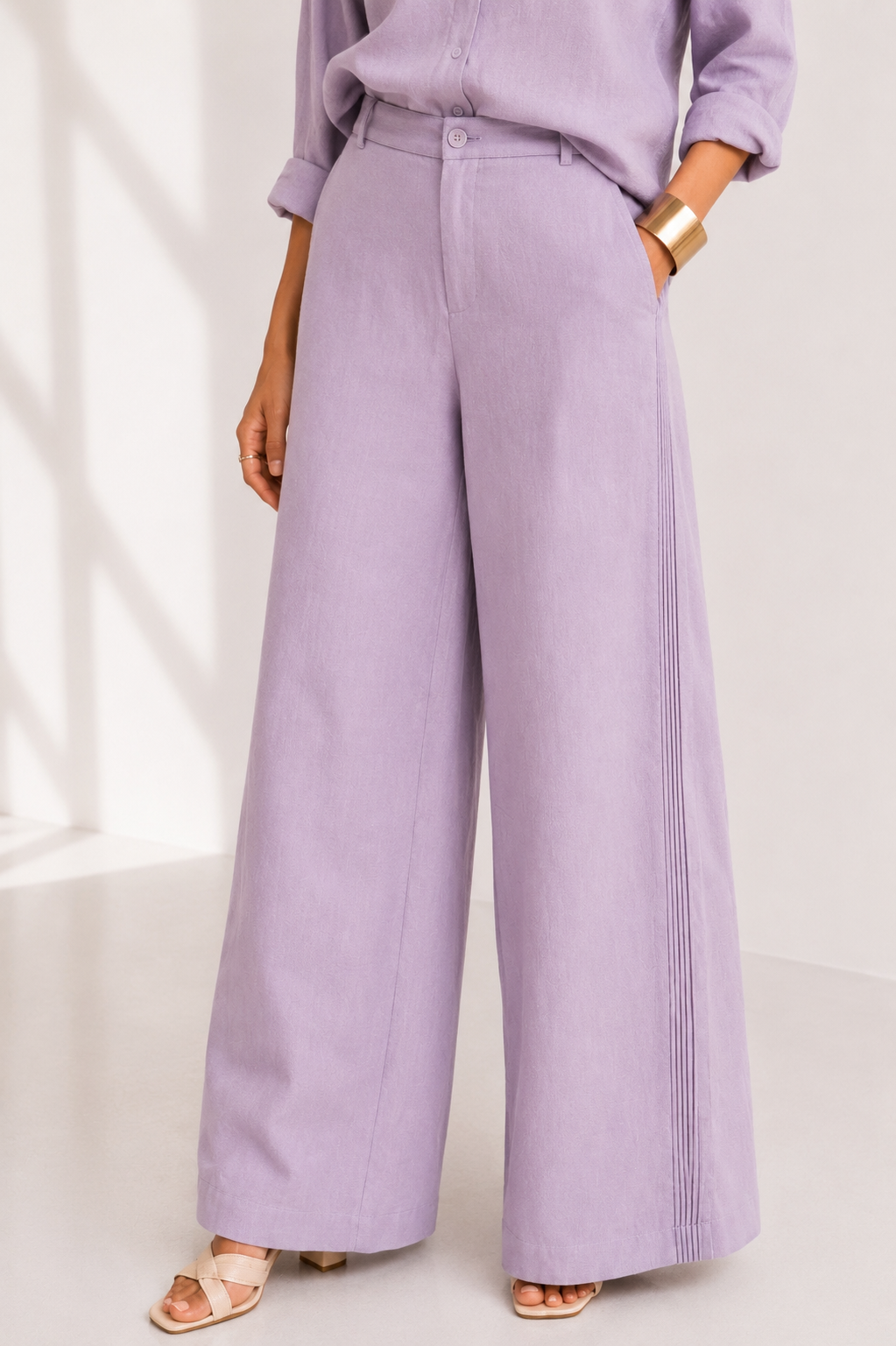 A person wearing wide-leg lavender pants with pleated side detailing, paired with a matching lavender top and beige sandals, set against a bright, sunlit studio background.